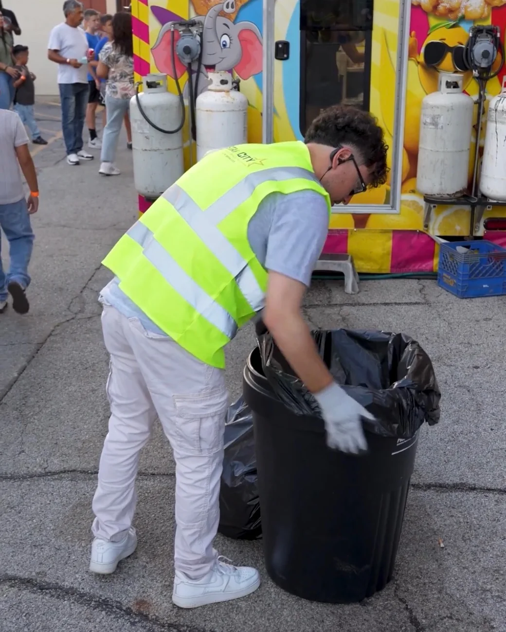 A crew member in a hi-vis vest bagging litter from a trash can in front of a funnel cake trailer