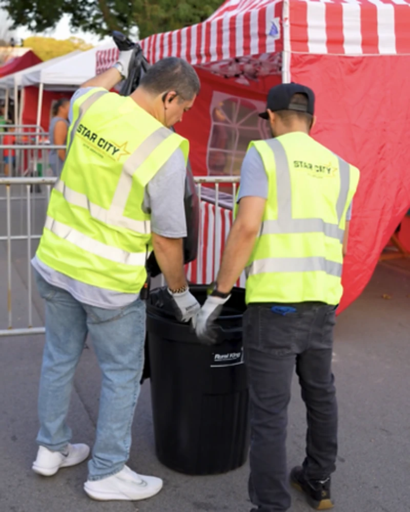 Two crew members in Star City Environmental vests lifting a bin liner out of a trash can beside a red and white striped tent