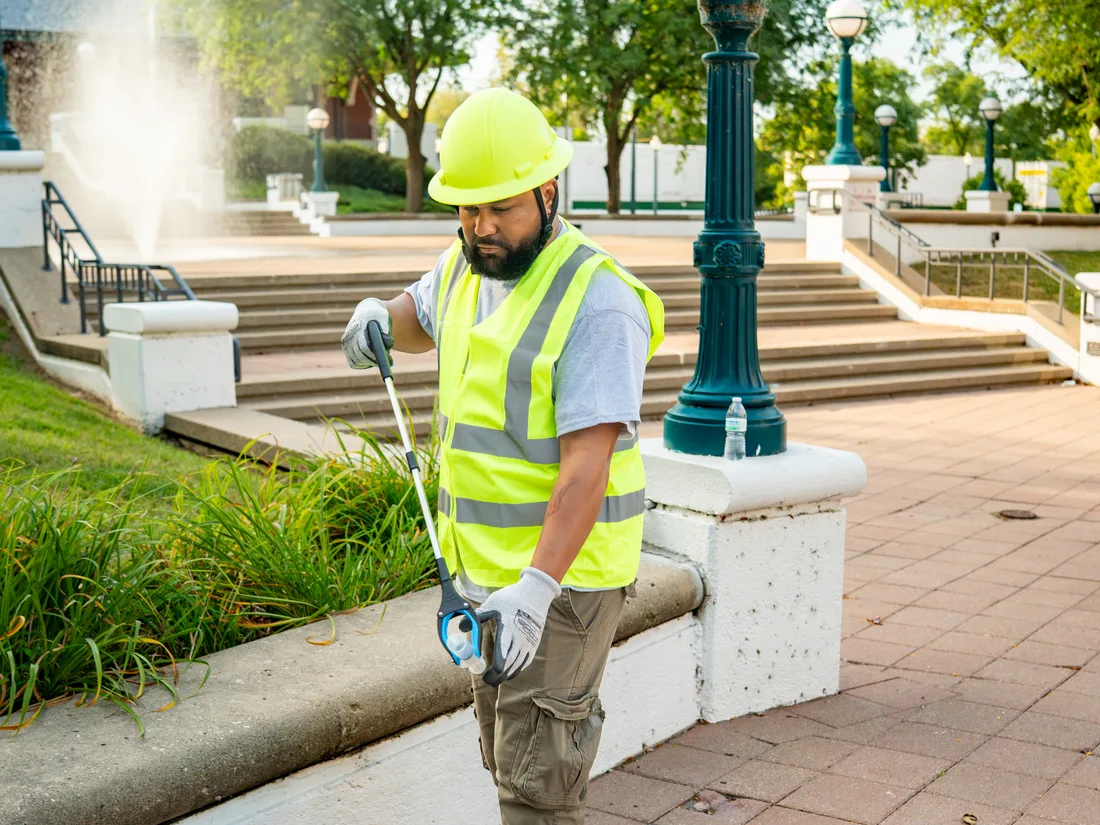 A Star City Environmental crew member in a hard hat, high-visibility vest and work gloves collecting litter on a public plaza.