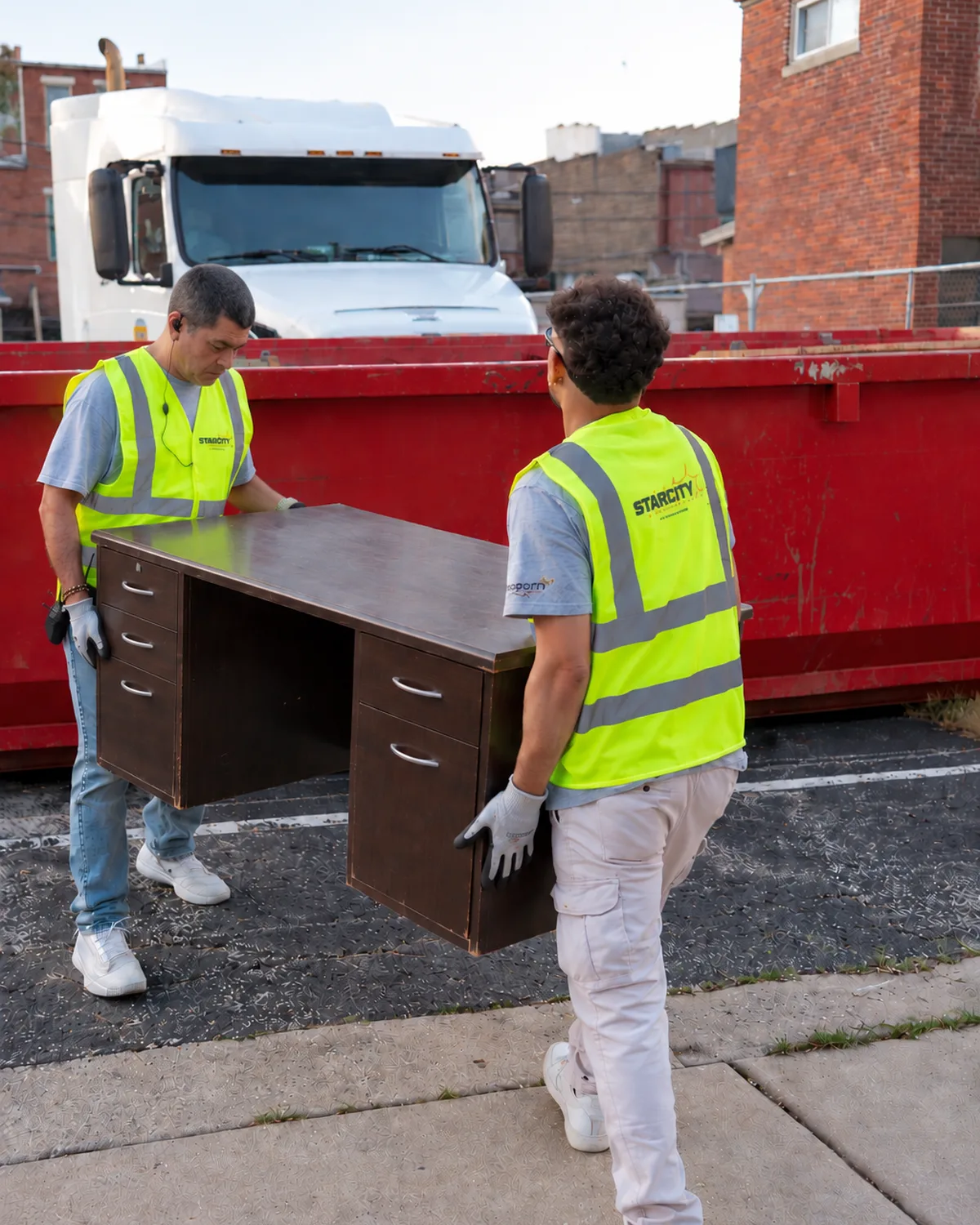 Two crew members in hi-vis vests lifting a wooden desk toward a red dumpster beside a truck.